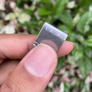 Może przedstawiać: Zbliżenie srebrnej klamry paska do zegarka z wygrawerowaną nazwą marki "CASIO". Klamra jest trzymana między palcami na tle zielonych i r&oacute;żowych liści.