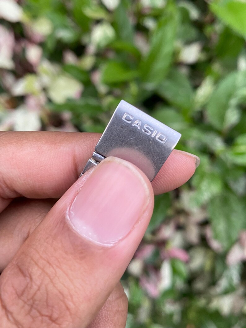 Może przedstawiać: Zbliżenie srebrnej klamry paska do zegarka z wygrawerowaną nazwą marki "CASIO". Klamra jest trzymana między palcami na tle zielonych i r&oacute;żowych liści.