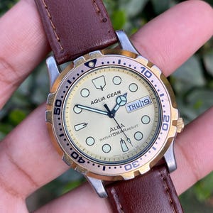 Pode incluir: Um relógio Aqua Gear vintage com uma pulseira de couro castanho. O mostrador creme tem detalhes dourados e exibe "THU 19". O relógio tem uma luneta rotativa e é resistente à água.