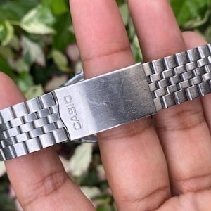 May include: A close-up of a silver-toned Casio watch band. The band is made of small, rectangular metal links. The clasp is visible, with the brand name "CASIO" engraved on it. The watch band is held by a person's fingers.