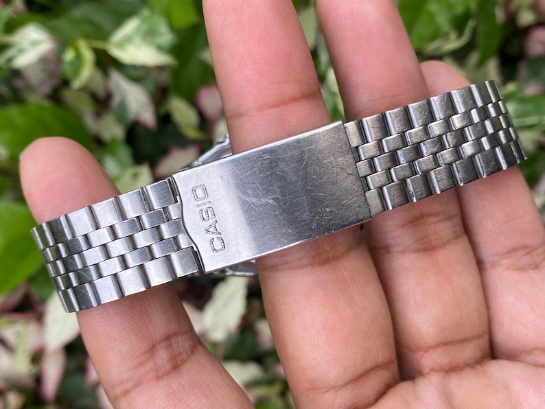 May include: A close-up of a silver-toned Casio watch band. The band is made of small, rectangular metal links. The clasp is visible, with the brand name "CASIO" engraved on it. The watch band is held by a person's fingers.