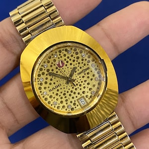 Könnte beinhalten: Goldfarbene Rado Diastar Uhr mit einem einzigartigen Zifferblatt mit gepunktetem Muster und kleinen Kristallen. Die Uhr hat ein goldenes Armband und eine Datumsanzeige bei 3 Uhr. Eine klassische Armbanduhr.