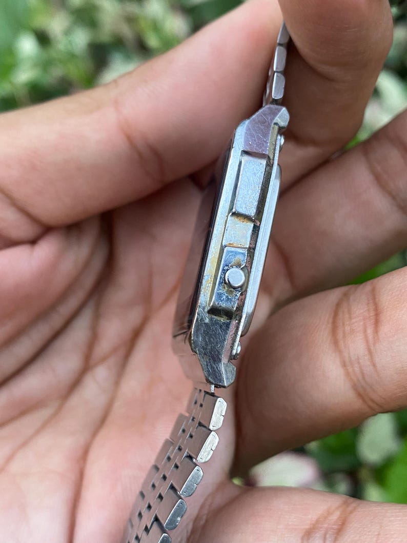 Może przedstawiać: Srebrny cyfrowy zegarek na rękę z prostokątną tarczą i metalową bransoletą z ogniwami. Zegarek nosi ślady użytkowania, z przebarwieniami z boku. Bransoleta składa się z małych, prostokątnych ogniw.