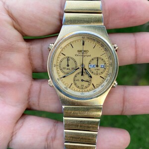 May include: A gold-tone Seiko Quartz chronograph watch with a round champagne-colored dial, three sub-dials, and a day-date window. The watch features a tachymeter scale on the outer bezel and a matching gold-tone metal link bracelet strap.
