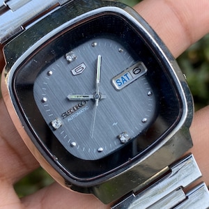Puede incluir: Un reloj Seiko 5 Automatic vintage con esfera gris cuadrada. El reloj presenta una correa y caja de acero inoxidable plateado. La esfera muestra el día y la fecha, con marcadores en forma de diamante. Las manecillas del reloj son plateadas.