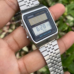 Może przedstawiać: Srebrny cyfrowy zegarek Casio z czarną tarczą wyświetlającą godzinę 6:28:15. Zegarek posiada napisy "CASIO", "ALARM CHRONO" i "WATER RESIST". Pasek zegarka wykonany jest z metalowych ogniw.