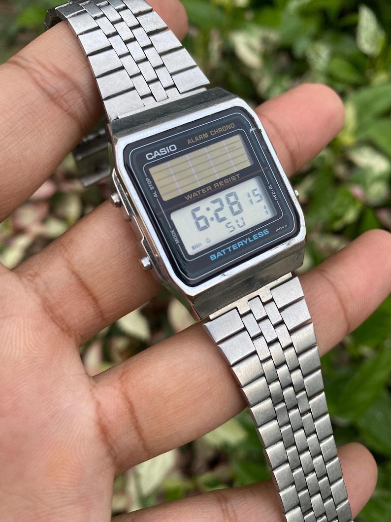Może przedstawiać: Srebrny cyfrowy zegarek Casio z czarną tarczą wyświetlającą godzinę 6:28:15. Zegarek posiada napisy "CASIO", "ALARM CHRONO" i "WATER RESIST". Pasek zegarka wykonany jest z metalowych ogniw.