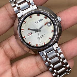 Puede incluir: Un reloj Citizen Crystal Seven vintage con esfera plateada y correa de acero inoxidable. La esfera muestra el día y la fecha, con detalles en rojo. Un reloj clásico.