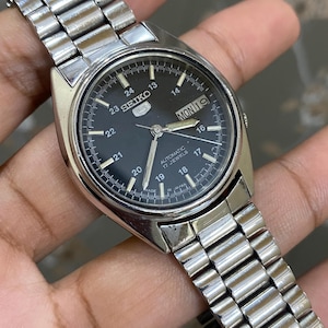 May include: A silver-tone stainless steel Seiko 5 automatic wristwatch with a black dial, featuring a day-date window at the three o'clock position, luminous hands, and a 24-hour military time scale printed in white on the watch face.