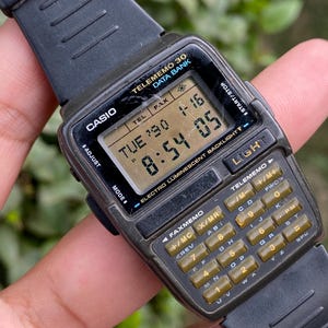 May include: A vintage Casio Telememo 30 Data Bank watch with a black resin strap. The watch face shows the time, date, and features a calculator keypad. The watch has the text "TELEMEMO 30 DATA BANK" and "ELECTRO LUMINESCENT BACKLIGHT".