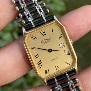 May include: A gold-toned Kolber Geneva quartz watch with a rectangular face and Roman numeral hour markers. The watch features a date display and a black and silver link band. The watch is a classic timepiece.