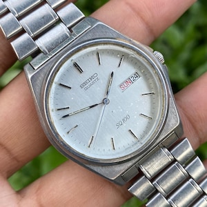 May include: A silver-toned Seiko Quartz SQ100 wristwatch with a metallic link band. The watch face is light silver with thin black hour markers, silver hands, and a rectangular day-date window at the three o'clock position displaying SUN 24.