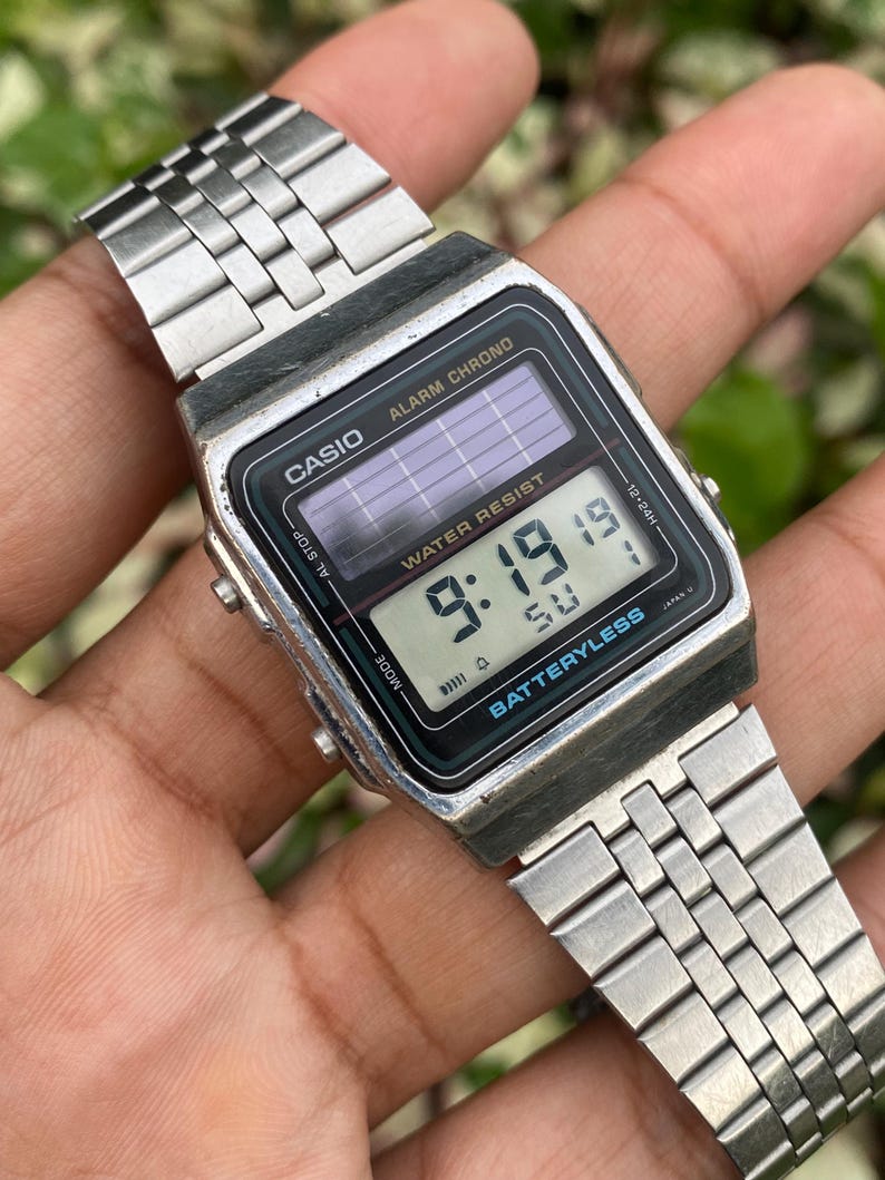 May include: A silver Casio digital watch with a black face displaying the time 9:19. The watch has a rectangular face, a metal band, and the text "CASIO" and "BATTERYLESS" are visible. The watch is a vintage style.