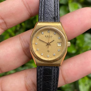 Könnte beinhalten: Eine goldfarbene Rado Quartz-Uhr mit einem schwarzen Lederarmband. Das Zifferblatt der Uhr ist goldfarben mit Diamant-Stundenmarkierungen und einer Datumsanzeige auf 3 Uhr. Die Uhr wird vor einem verschwommenen grünen Hintergrund gehalten.