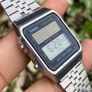 Może przedstawiać: Srebrny cyfrowy zegarek Casio z czarną tarczą wskazującą godzinę 6:28:40. Zegarek ma prostokątną tarczę z napisami "CASIO", "ALARM CHRONO" i "WATER RESIST". Pasek zegarka jest wykonany ze srebrnego metalu.