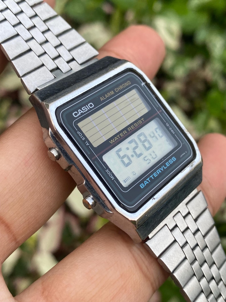 Może przedstawiać: Srebrny cyfrowy zegarek Casio z czarną tarczą wskazującą godzinę 6:28:40. Zegarek ma prostokątną tarczę z napisami "CASIO", "ALARM CHRONO" i "WATER RESIST". Pasek zegarka jest wykonany ze srebrnego metalu.