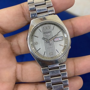 May include: Silver-toned Seiko 5 automatic watch with a rectangular face. The watch features a silver dial with vertical stripes, a day-date display, and a stainless steel band. The watch is shown against a blue background.