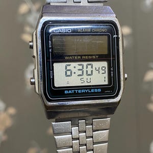 Może przedstawiać: Srebrny cyfrowy zegarek Casio pokazujący godzinę 6:30:49 w niedzielę. Tarcza zegarka ma czarną obw&oacute;dkę z napisami "CASIO ALARM CHRONO" i "WATER RESIST". Pasek zegarka wykonany jest ze srebrnych metalowych ogniw.