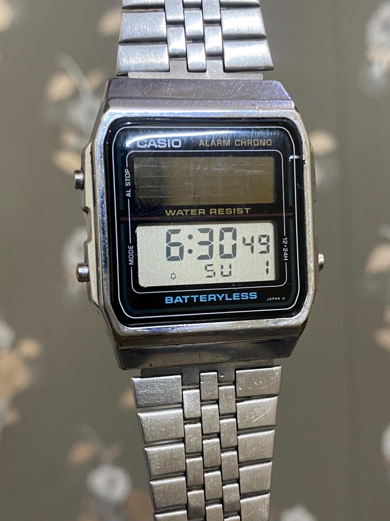 Może przedstawiać: Srebrny cyfrowy zegarek Casio pokazujący godzinę 6:30:49 w niedzielę. Tarcza zegarka ma czarną obw&oacute;dkę z napisami "CASIO ALARM CHRONO" i "WATER RESIST". Pasek zegarka wykonany jest ze srebrnych metalowych ogniw.