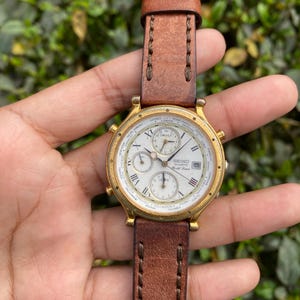 May include: A gold-toned Seiko Quartz watch with a white face, featuring multiple subdials and Roman numeral hour markers. The watch has a brown leather strap. The watch face displays the text "SEIKO Quartz World Time".