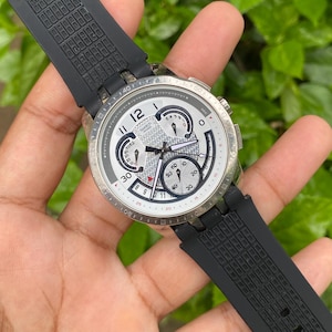 May include: A silver-toned wristwatch with a white face, black hands, and a black rubber strap. The watch face features multiple sub-dials and the number 12 in a bold font. The watch is held by a person.