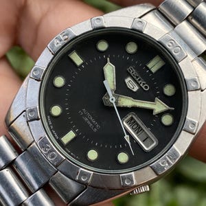 May include: A stainless steel Seiko automatic watch with a black dial. The watch has luminous hands and markers, a day-date display, and a rotating bezel. The watch band is also stainless steel.