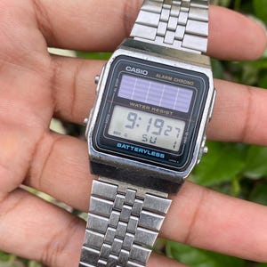 May include: A silver Casio digital watch with a rectangular black face displaying the time 9:19:27. The watch has a silver metal band and the text "WATER RESIST" and "BATTERYLESS" are visible on the face.