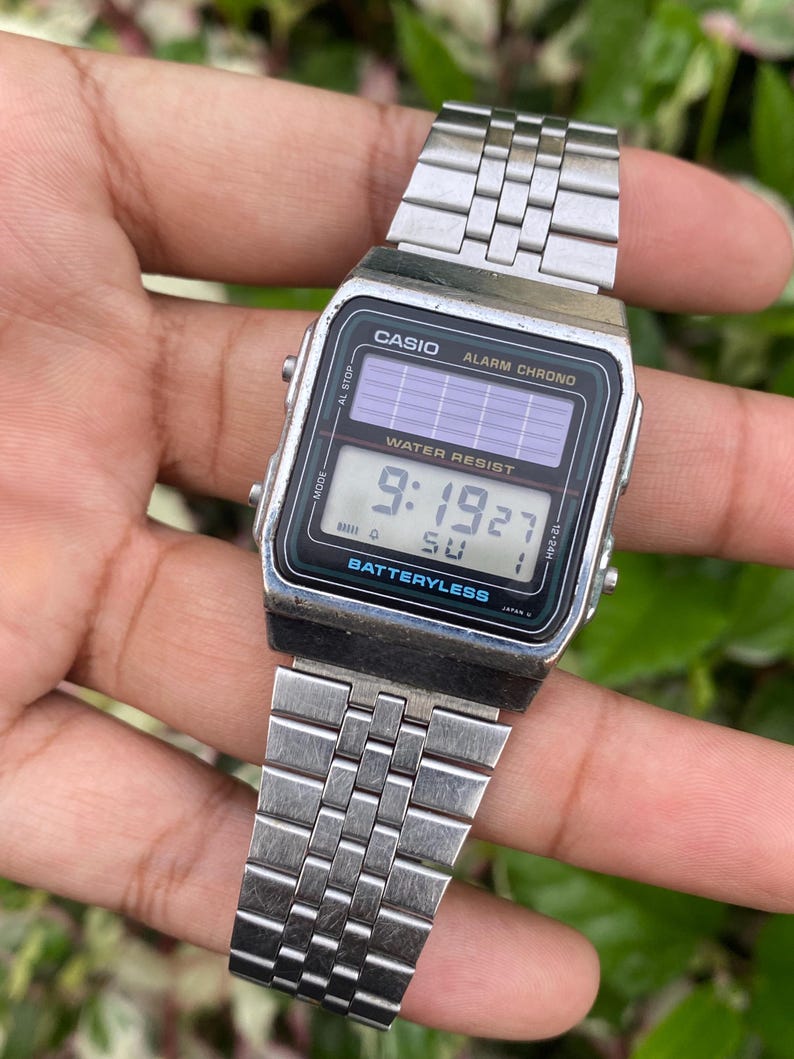 May include: A silver Casio digital watch with a rectangular black face displaying the time 9:19:27. The watch has a silver metal band and the text "WATER RESIST" and "BATTERYLESS" are visible on the face.