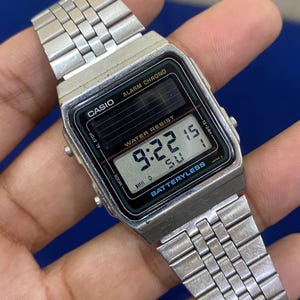 May include: A silver Casio digital watch with a rectangular black face displaying the time 9:22. The watch has a silver band and features the text "ALARM CHRONO" and "WATER RESIST". The watch is battery-less.