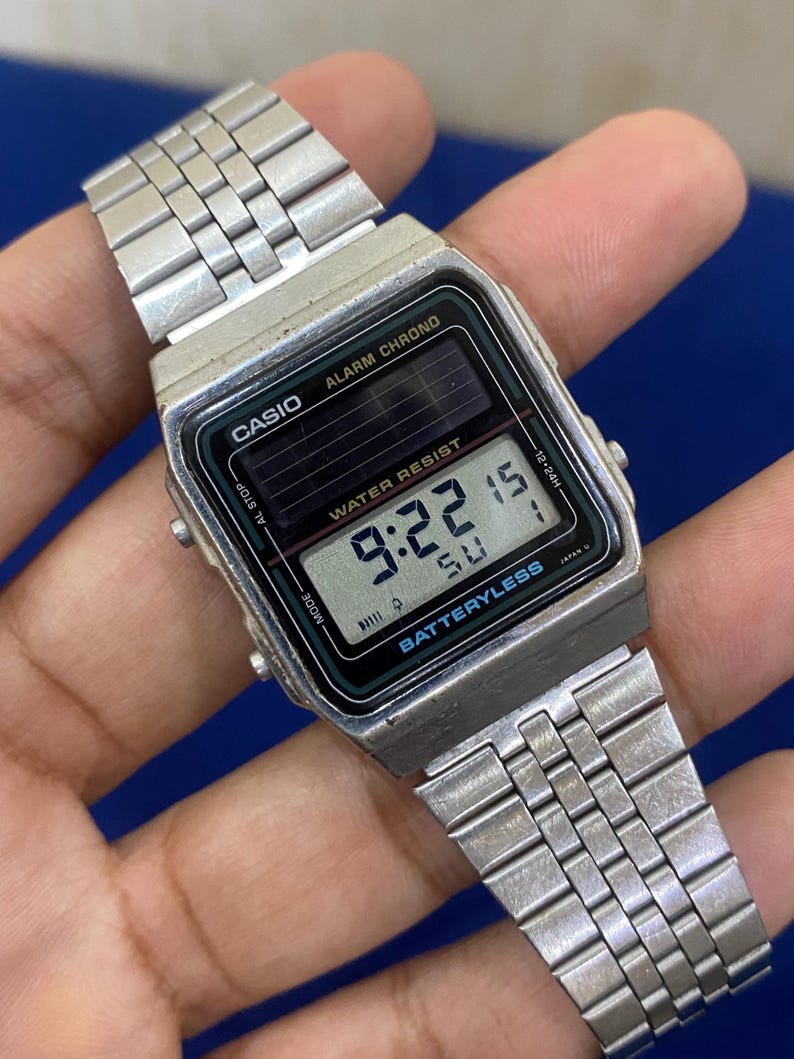 May include: A silver Casio digital watch with a rectangular black face displaying the time 9:22. The watch has a silver band and features the text "ALARM CHRONO" and "WATER RESIST". The watch is battery-less.