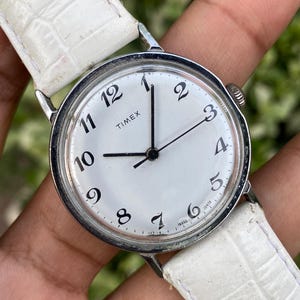 Könnte beinhalten: Eine Timex Armbanduhr mit rundem silberfarbenem Gehäuse und weißem Zifferblatt mit schwarzen arabischen Ziffern. Die Uhr hat schwarze Stunden-, Minuten- und Sekundenzeiger und ist mit einem weißen, strukturierten Lederarmband versehen.