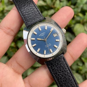 Pode incluir: Um relógio de pulso Rotary vintage com mostrador azul e ponteiros prateados. O relógio apresenta uma janela de data às 3 horas e uma bracelete de couro preta. O mostrador exibe o texto "Rotary" e "21 Jewels Automatic".