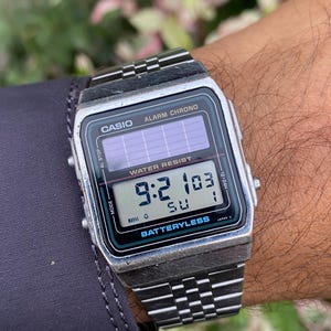 May include: A silver Casio digital watch with a black face displaying the time 9:21:03. The watch has a rectangular face with the words "CASIO", "ALARM CHRONO", and "WATER RESIST" printed on it. The watch band is silver.