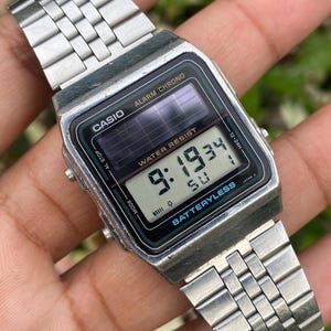 May include: A vintage Casio digital watch with a silver-tone metal band. The watch face displays the time, date, and other functions. The watch has the text "CASIO", "ALARM CHRONO", "WATER RESIST", and "BATTERYLESS".