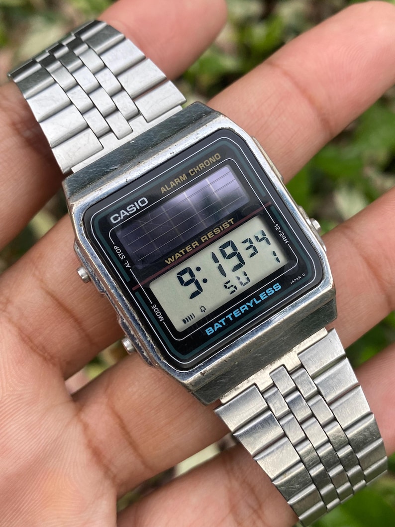 May include: A vintage Casio digital watch with a silver-tone metal band. The watch face displays the time, date, and other functions. The watch has the text "CASIO", "ALARM CHRONO", "WATER RESIST", and "BATTERYLESS".