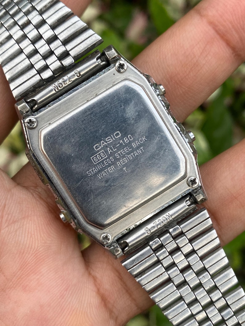 Może przedstawiać: Srebrny zegarek cyfrowy Casio AL-180, widok z tyłu. Tył ze stali nierdzewnej zawiera logo Casio i informacje o modelu. Pasek zegarka ma srebrny, połączony design.