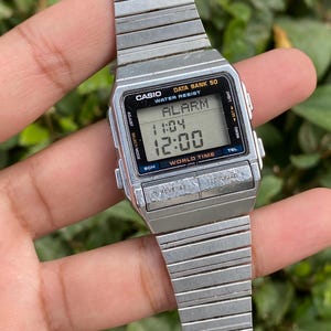 Può includere: Un orologio digitale Casio Data Bank 50 argentato con un quadrante rettangolare che mostra l'ora e le impostazioni dell'allarme. L'orologio ha un cinturino in metallo e diversi pulsanti per regolare le impostazioni. Il quadrante dell'orologio indica "ALARM" e "WATER RESIST".