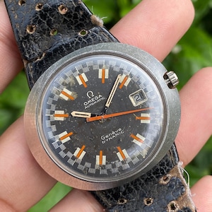 Peut inclure: Montre automatique Omega Genève Dynamic vintage avec un cadran noir, des marqueurs orange et blancs, et une fenêtre de date à 3 heures. La montre a un boîtier en acier inoxydable et un bracelet en cuir noir.