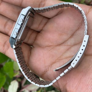 May include: A silver-toned digital watch with a rectangular face and a matching metal band. The watch has two buttons on the side and a clasp closure. The watch is a classic style.