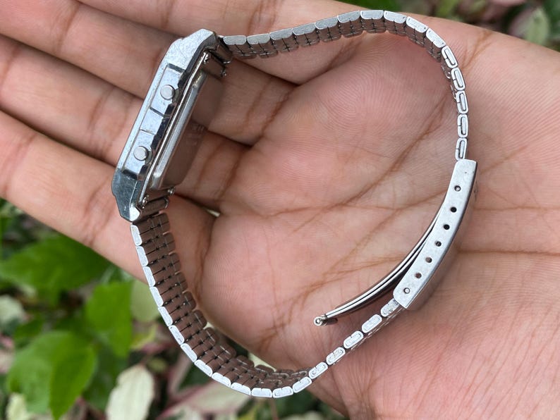 May include: A silver-toned digital watch with a rectangular face and a matching metal band. The watch has two buttons on the side and a clasp closure. The watch is a classic style.