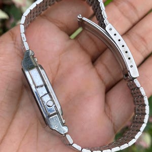 May include: A silver-toned wristwatch with a rectangular face and a linked metal band. The watch is shown from a side angle, displaying the clasp and the details of the band's construction. The watch appears to be vintage or retro in style.