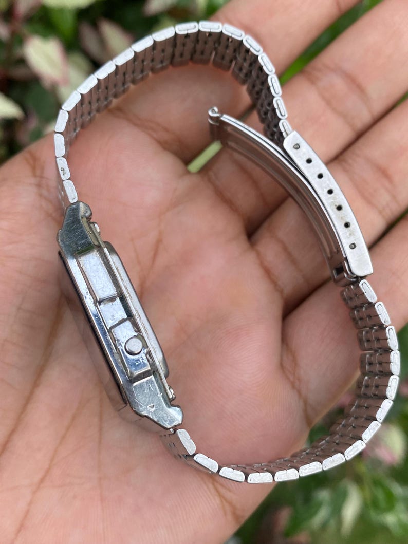May include: A silver-toned wristwatch with a rectangular face and a linked metal band. The watch is shown from a side angle, displaying the clasp and the details of the band's construction. The watch appears to be vintage or retro in style.