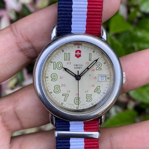 May include: A Swiss Army watch with a cream-colored face, silver case, and a red cross logo. The watch has a nylon band with blue, white, and red stripes. The watch face displays the numbers 1 through 12 and 13 through 23.
