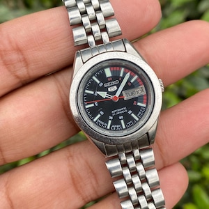 May include: A vintage Seiko 5 automatic wristwatch with a black dial and silver-tone stainless steel band. The watch face displays the day and date, with red accents. The watch is held in a hand against a blurred green background.