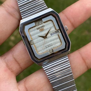 Op de afbeelding: Een zilverkleurig Seiko 5 automatisch horloge met een rechthoekige kast en een getextureerde witte wijzerplaat. Het horloge heeft goudkleurige wijzers, een dag-datumvenster op de drie-uurpositie en vier kleine heldere steentjes op de hoeken. Het heeft een flexibele metalen rekband.
