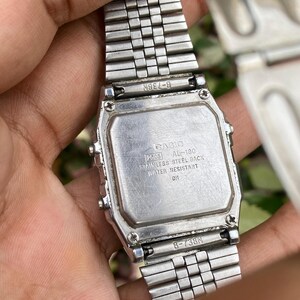 May include: A silver Casio digital watch with a stainless steel back. The watch band is also silver and made of small, connected metal pieces. The watch face has the text "CASIO 2505 AL-180 STAINLESS STEEL BACK WATER RESISTANT DH".