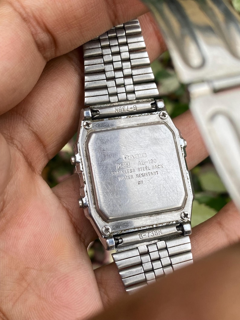 May include: A silver Casio digital watch with a stainless steel back. The watch band is also silver and made of small, connected metal pieces. The watch face has the text "CASIO 2505 AL-180 STAINLESS STEEL BACK WATER RESISTANT DH".