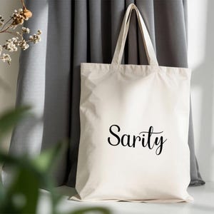 May include: Cream-colored tote bag with the word "Sarity" in black script. The bag has two handles and is made of a sturdy fabric. The background includes a gray curtain and a hint of greenery.