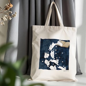 May include: Cream-colored tote bag featuring a dark blue square with white rabbits, stars, and an open book illustration. The bag has long handles and is made of a sturdy fabric, perfect for carrying books or groceries.