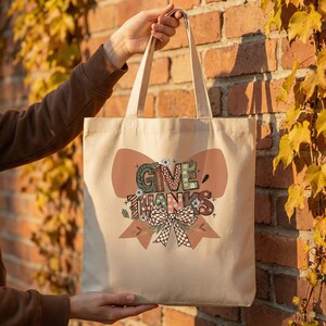 Könnte beinhalten: Beige Tragetasche mit großem Schleifendesign und den Worten "GIVE THANKS" in dekorativer Schrift. Der Bogen weist eine Mischung aus Farben wie Braun, Grün und Weiß mit floralen und karierten Mustern auf. Die Tasche wird von einer Person gehalten.